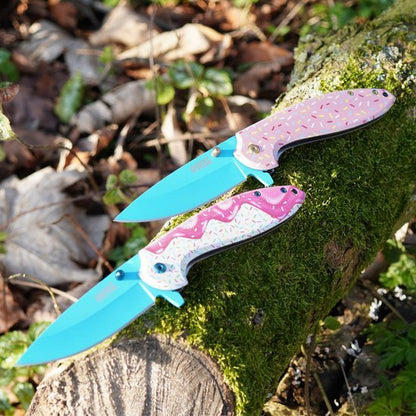 Twee kleurrijke opvouwbare messen met blauw lemmet en roze handvat met glazuur en sprinkels, op mos.